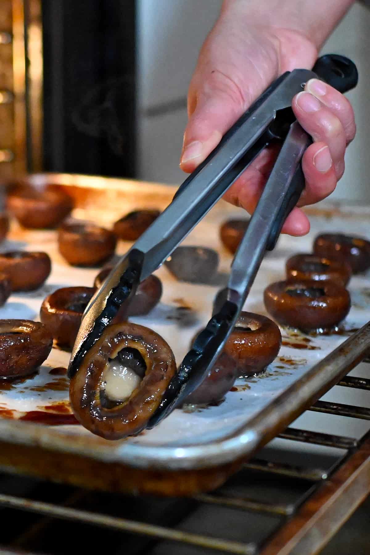 A pair of tongs are flipping over the roasted cremini mushrooms so they are gill-side up.