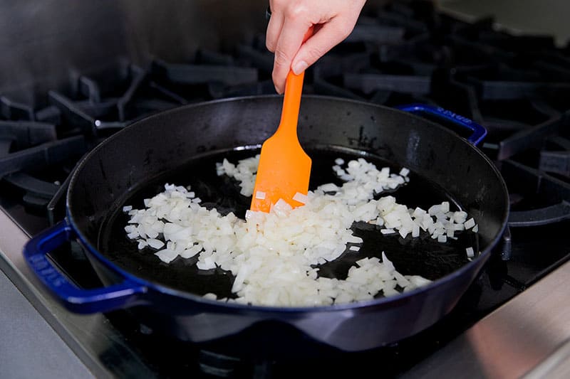 A orange silicone spatula is stirring diced onions in a cast iron skillet
