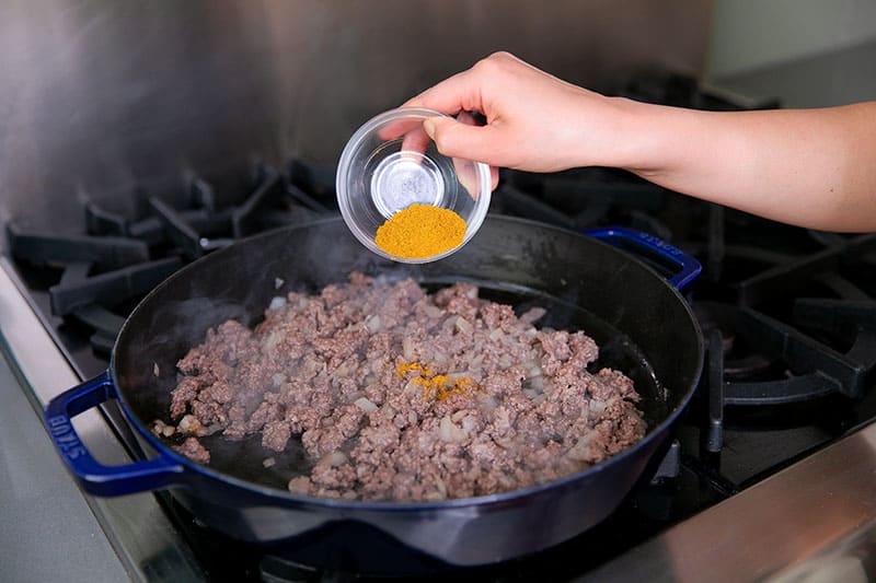 Pouring a small glass bowl filled with Indian curry powder on a cast iron skillet filled with ground beef and onions.