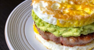 A white plate topped with a paleo egg McMuffin breakfast sandwich made with two fried egg buns and a sausage patty filling and guacamole.