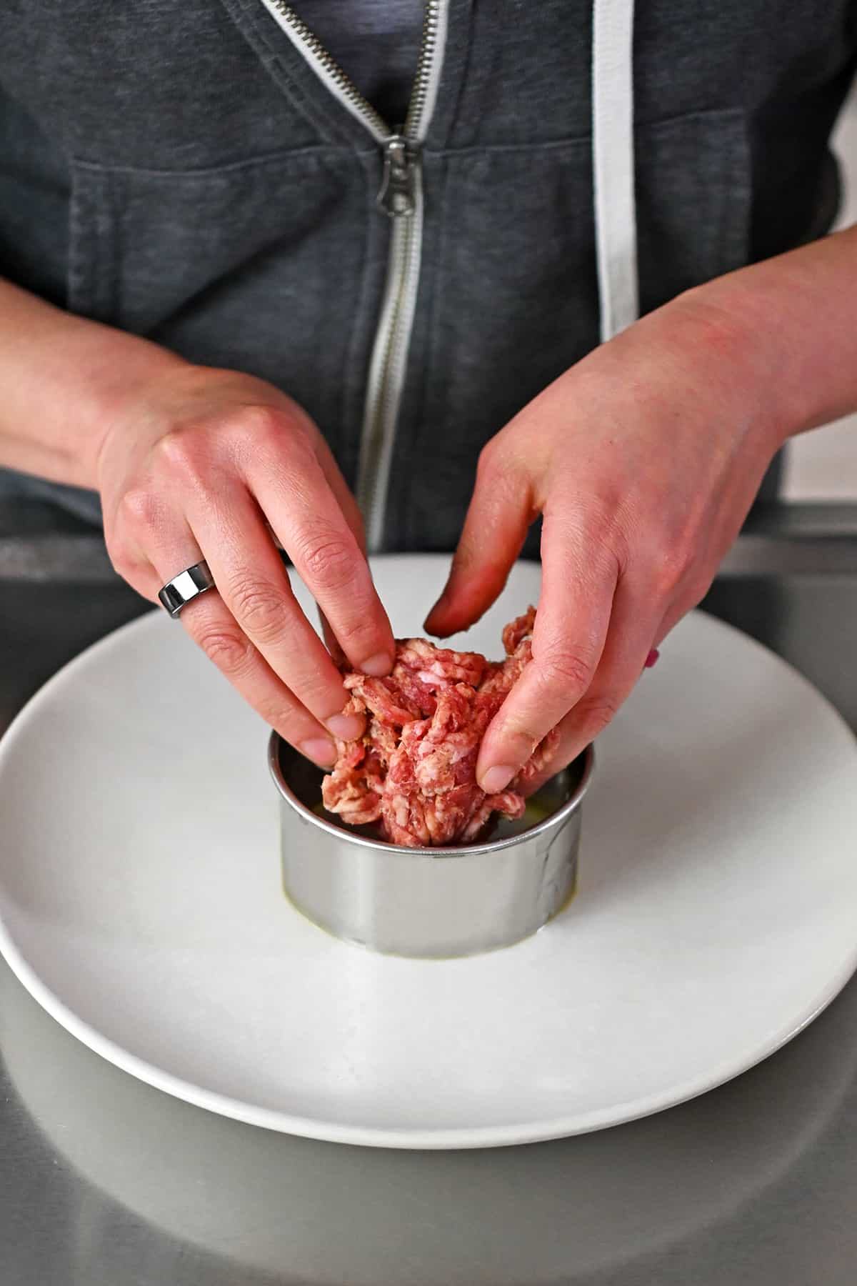 Two hands are adding raw bulk sausage into a round metal biscuit cutter on a white plate.