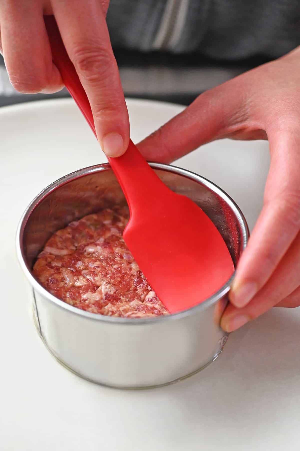 A red spatula is pressing raw bulk sausage into a stainless steel round biscuit mold.