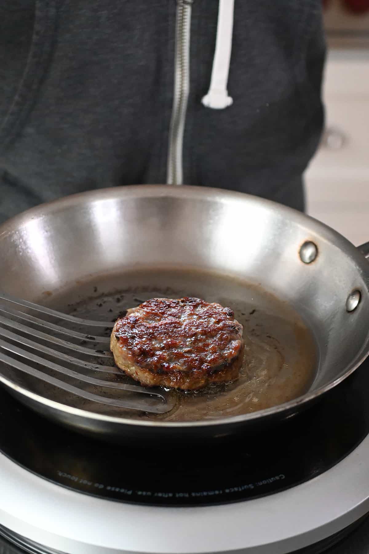 A fish spatula is removing a golden brown round sausage patty off of a frying pan.