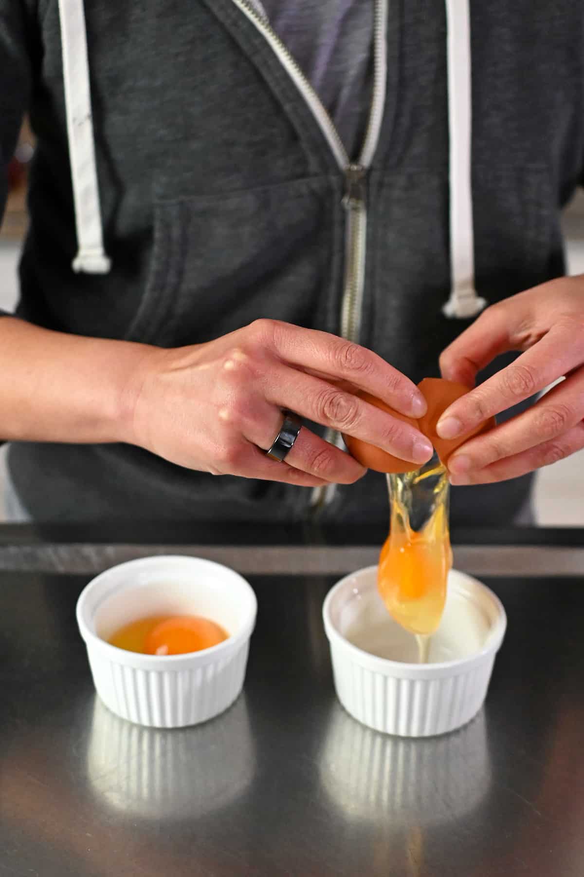 A person is cracking an egg into a white ramekin next to a ramekin containing a raw egg.