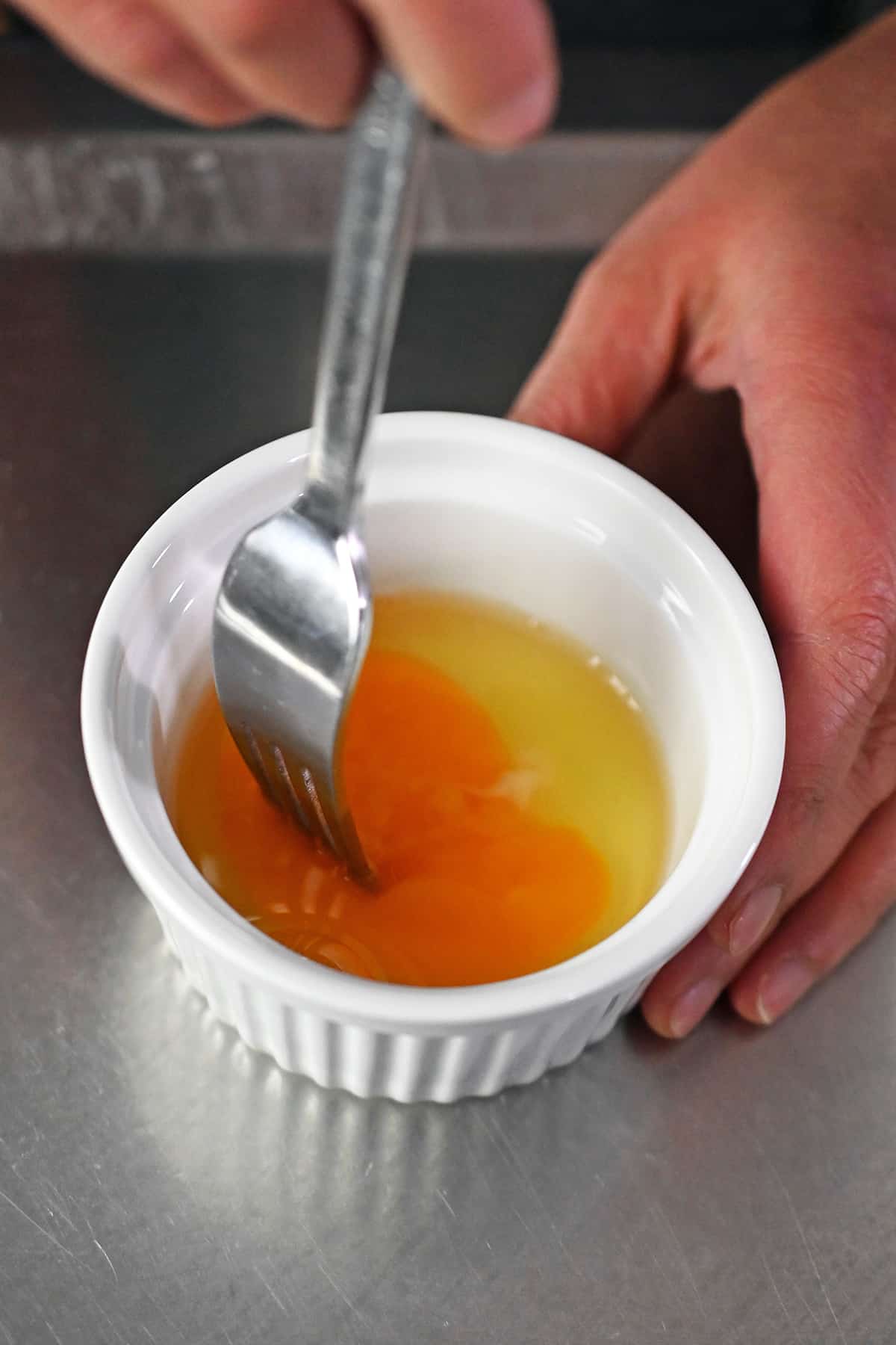 A closeup of a fork piercing the yolk of a raw egg in a white ramekin.