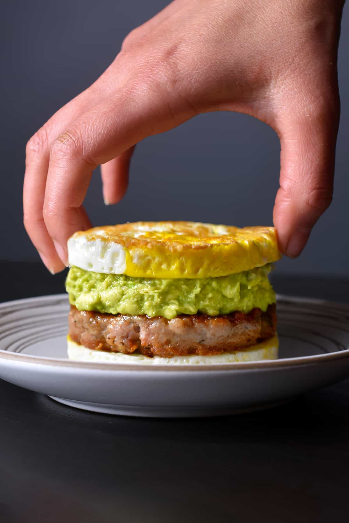 A hand is placing a fried egg on top of a sausage patty with guacamole.