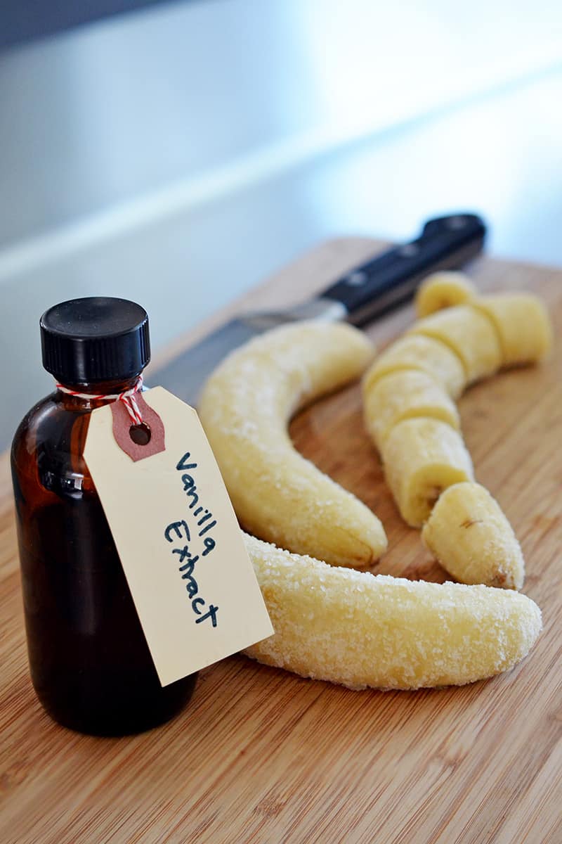Three frozen bananas and a bottle of vanilla extract are on a wooden cutting board.