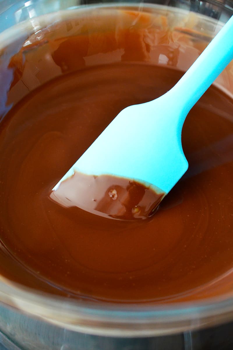 A blue silicone spatula is mixing a bowl of melted chocolate.