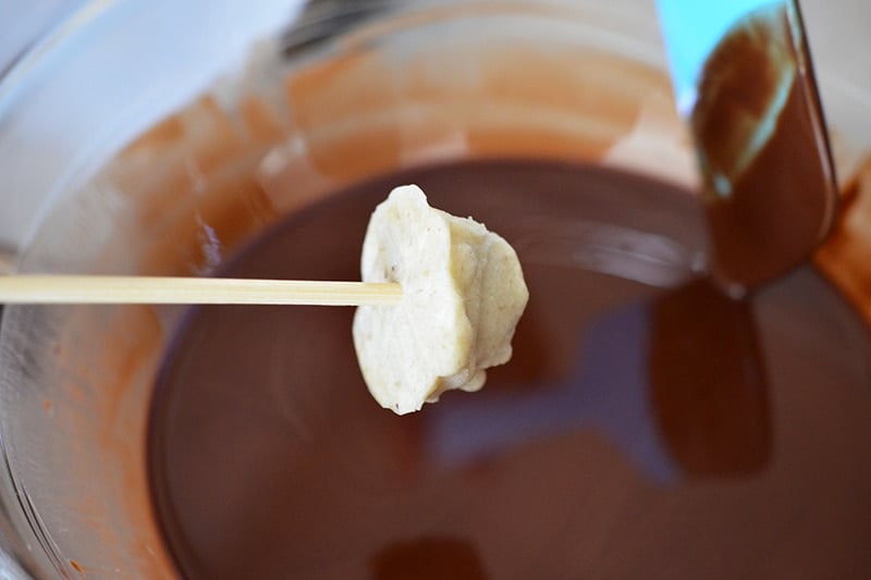 A banana ice cream scoop on a skewer is being dipped in melted chocolate to make Chunky Monkey Ice Cream Bon Bons.