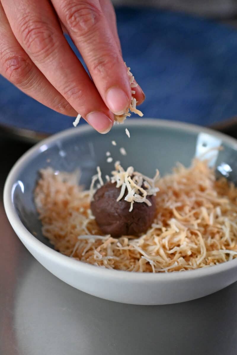 A hand is sprinkling toasted shredded coconut onto a paleo and vegan chocolate truffle in a small bowl.