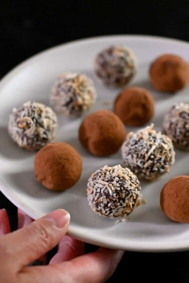 A hand is holding a white plate with paleo and vegan chocolate truffles topped with cocoa powder or toasted shredded coconut.