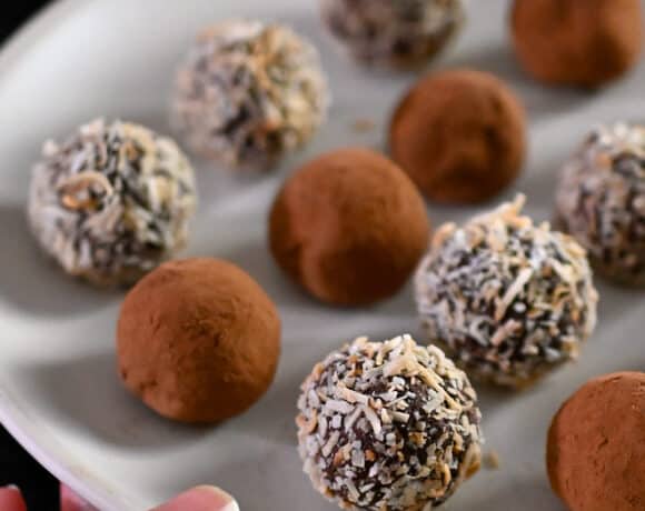 A hand is holding a white plate with paleo and vegan chocolate truffles topped with cocoa powder or toasted shredded coconut.