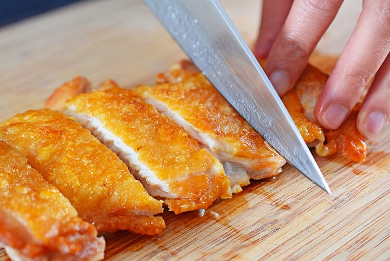 Slicing a piece of Cracklin' Chicken with a chef's knife on a wooden cutting board.