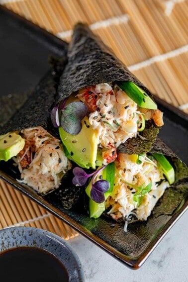 Three gluten-free and Whole30 California hand rolls stacked on a black plate, next to a dipping bowl with coconut aminos.