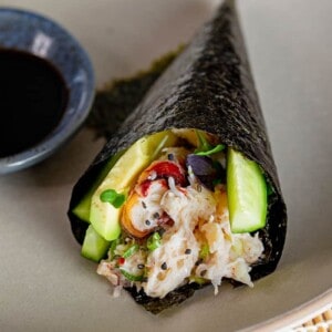 A closeup of a California hand roll on a gray plate next to a small bowl filled with coconut aminos.