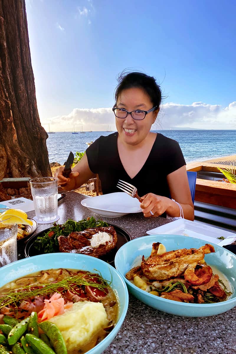 A smiling Asian woman is eating a gluten-free dinner by the beach in Maui