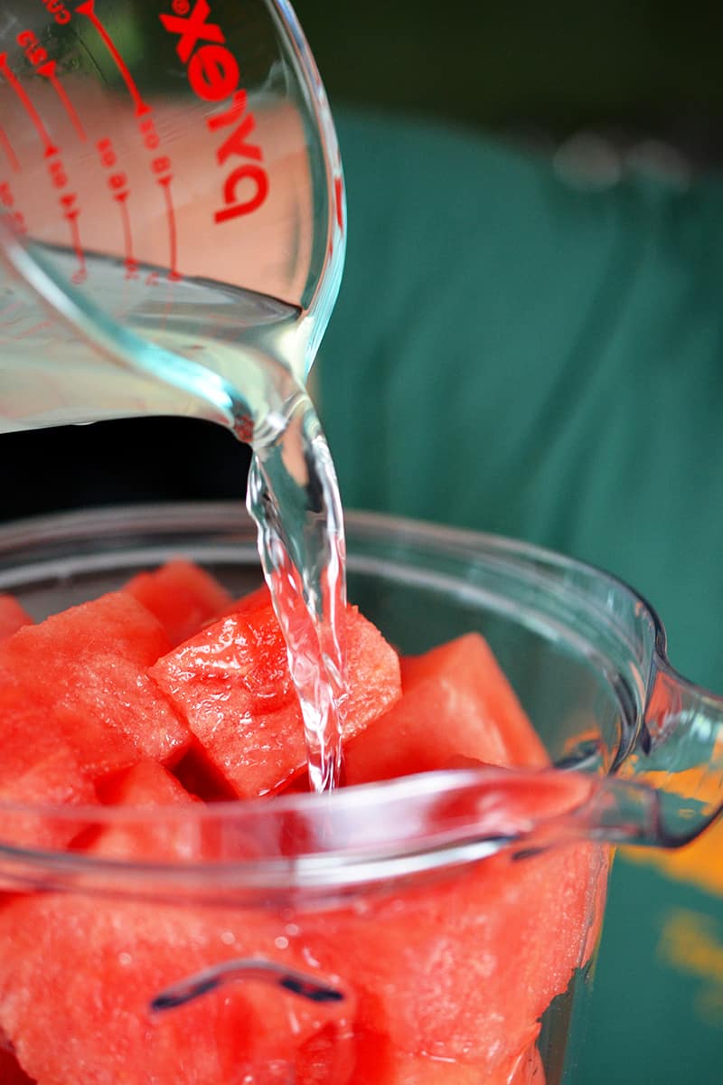 Pouring coconut water into a blender filled with watermelon cubes.