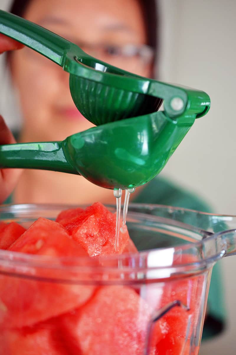 Someone using a green citrus juicer to add fresh lime juice to a blender filled with watermelon chunks