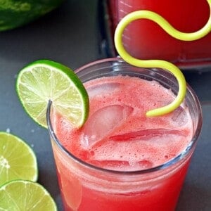 An overhead shot of watermelon juice cooler in a clear cup with lime wedges and a yellow straw.
