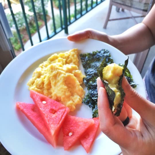 A hand is holding a white plate filled with watermelon slices, scrambled eggs, and toasted seaweed.