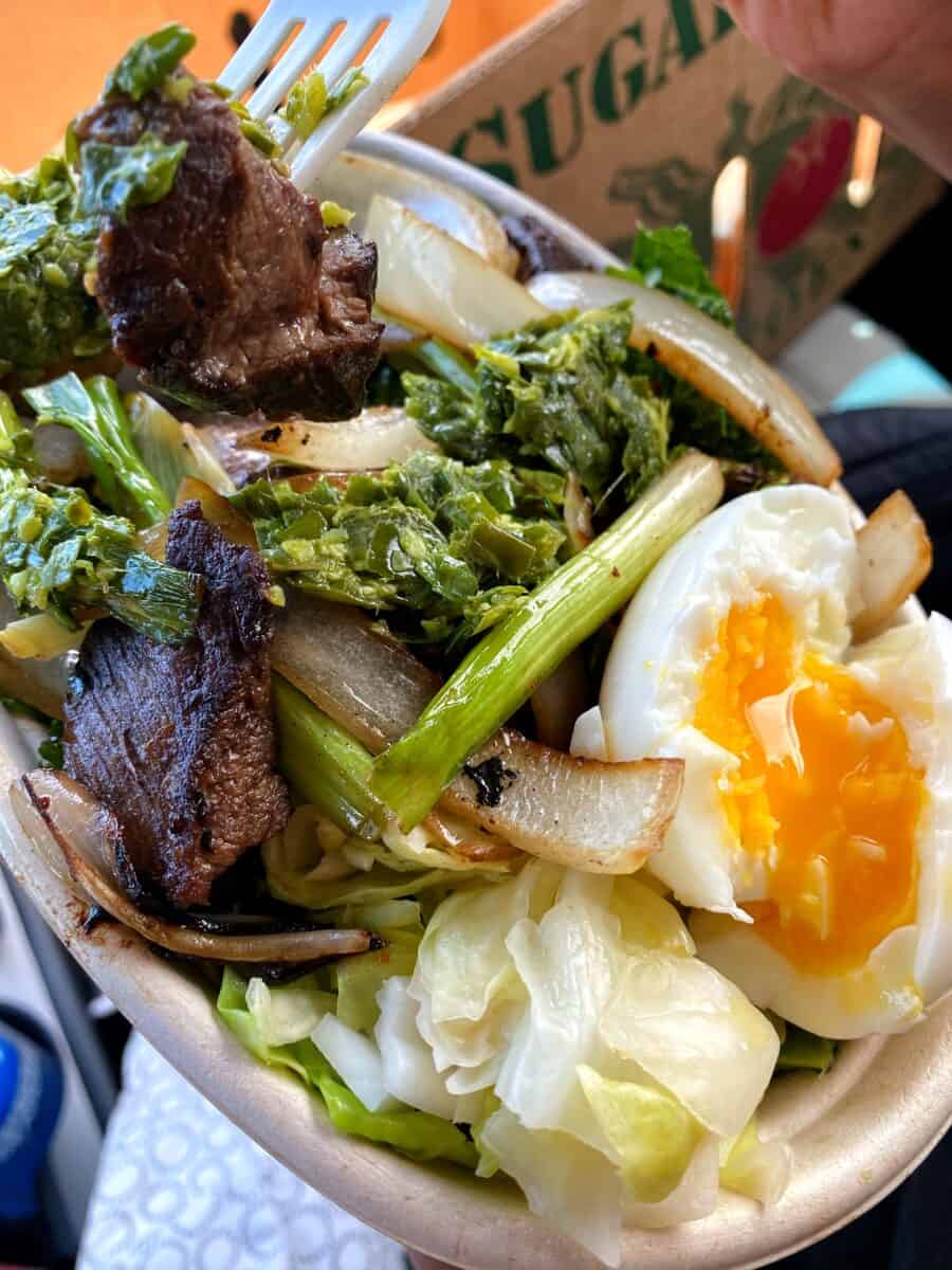 An overhead shot of a takeout container filled with cube steak, 6-minute egg, and kale salad from Tin Roof Maui