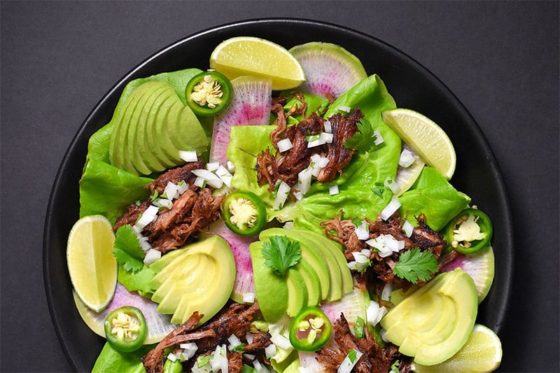 An overhead shot of Instant Pot Carnitas on a bed of lettuce and topped with sliced avocados, peppers, and herbs.