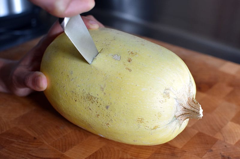 A small spaghetti squash is lying on it's side on a cutting board. There's a small paring knife stuck in the middle of the spaghetti squash, cutting it in half horizontally.