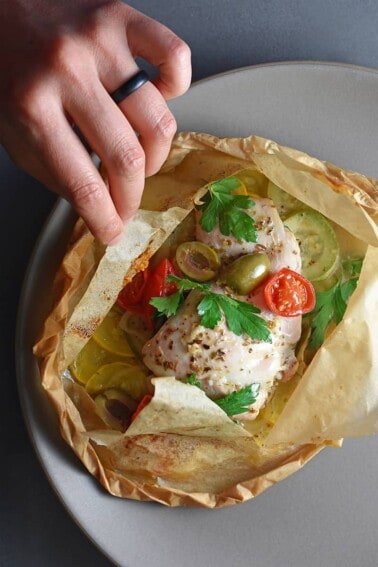 An overhead shot of an open lemon garlic chicken parchment packet filled with cooked chicken, olives, cherry tomatoes, and Italian parsley.