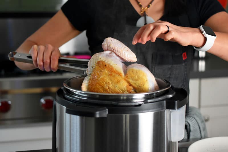 A person using tongs to turn a whole chicken that has been browning in an Instant Pot.