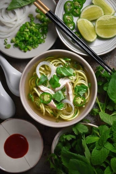 An overhead shot of a bowl of Instant Pot (Pressure Cooker) Chicken Pho surrounded by herbs, condiments, and sliced limes.