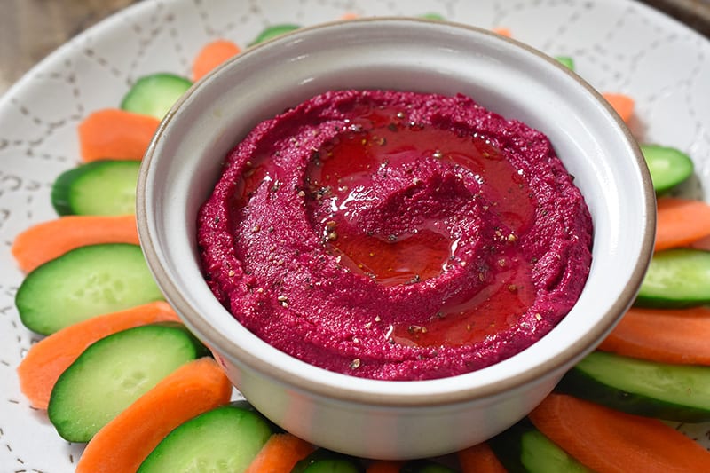 A bowl of paleo beet hummus in a plate with sliced carrots and cucumbers.