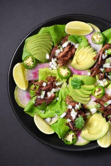 An overhead shot of Instant Pot (Pressure Cooker) Carnitas on a bed of lettuce with sliced peppers, avocado, and radishes.