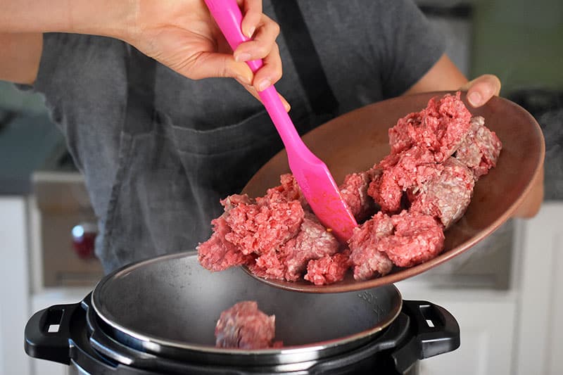 A woman is adding ground beef from a plate into an open Instant Pot.