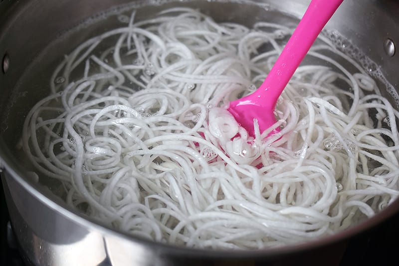 Daikon noodles cooking in a pot of water with a pink spatula inside the pot.