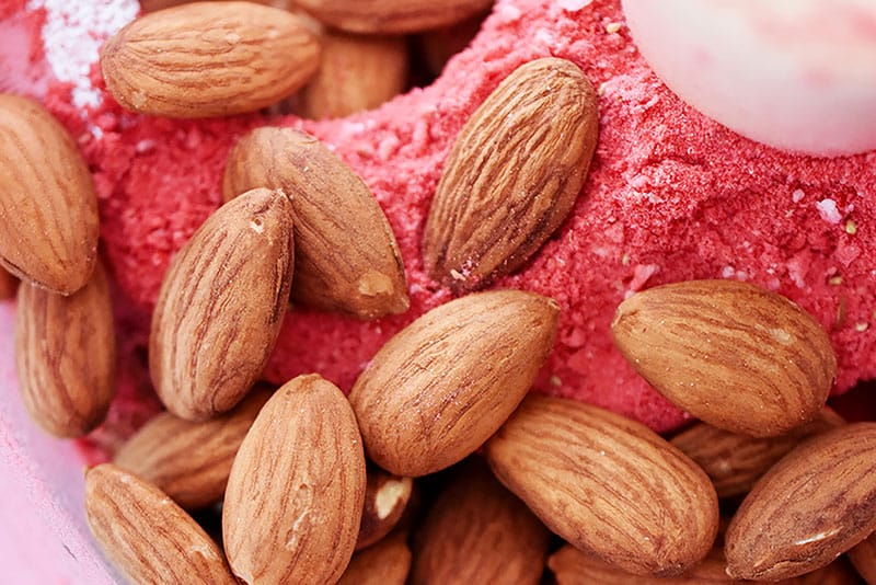 A closeup of toasted whole almonds on berry powder in a food processor.