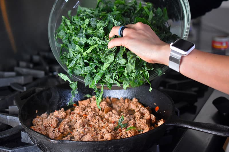 Shredded kale is added to the cooked Italian sausage in a large cast iron skillet.