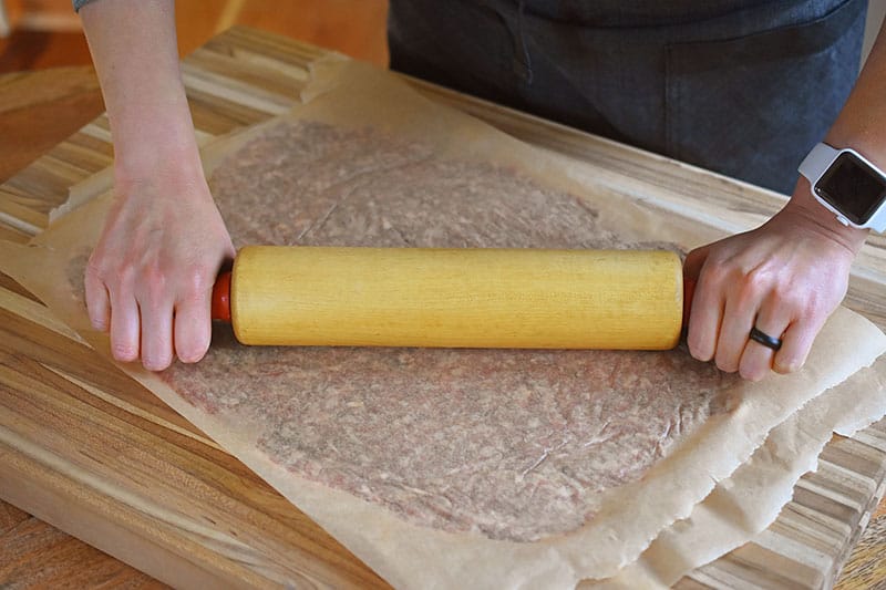 A meatza crust sandwiched between two pieces of parchment paper is being rolled flat with a rolling pin.