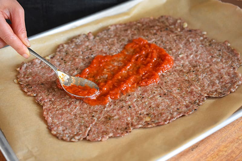 Spreading tomato sauce on the sausage crust of a meatza.