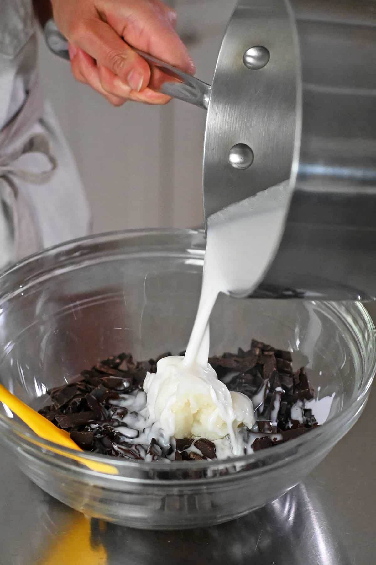 Pouring warm coconut milk into a bowl filled with dark chocolate slivers and coconut oil.