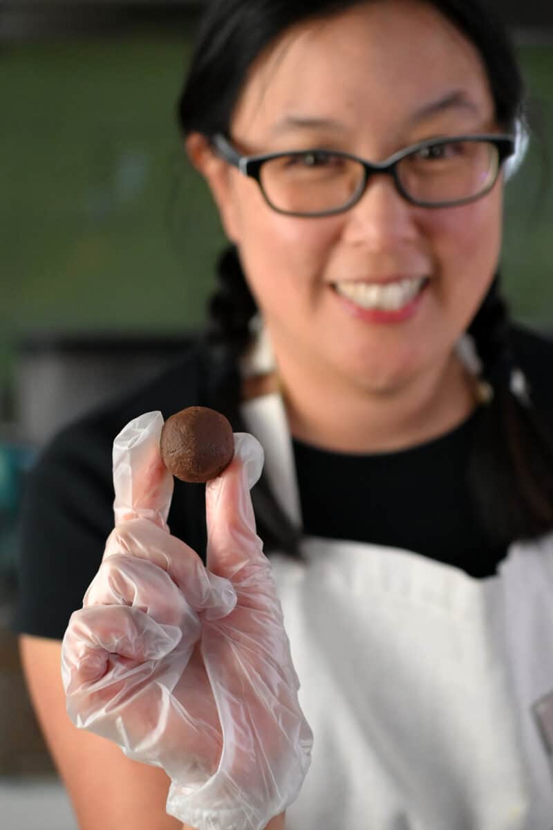 A smiling Asian woman in glasses is holding a round chocolate truffle in her gloved hand.