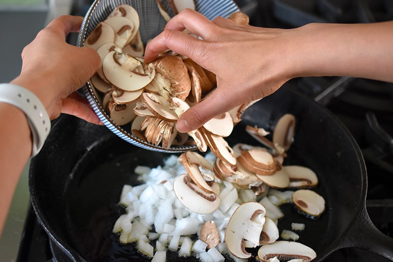 Adding diced onions and sliced mushrooms to a cast iron skillet.