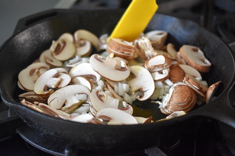 A yellow spatula stirs mushrooms and onions in a large cast iron skillet.