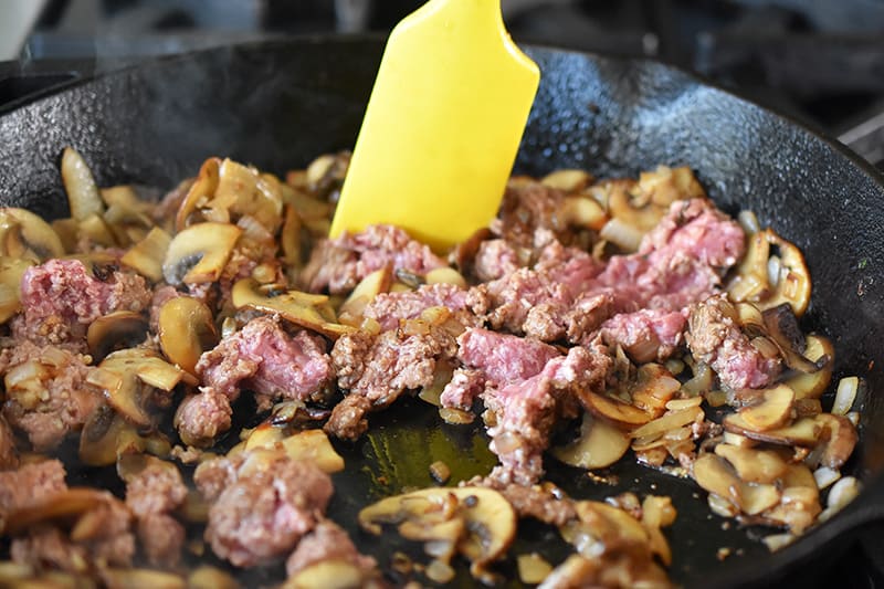 Ground beef is stirred into the pan with mushrooms, onions, and minced garlic.