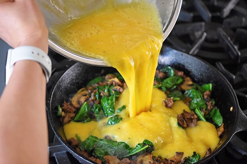 A bowl of scrambled eggs is poured into the pan with the cooked ground beef, spinach, mushrooms, and onion.