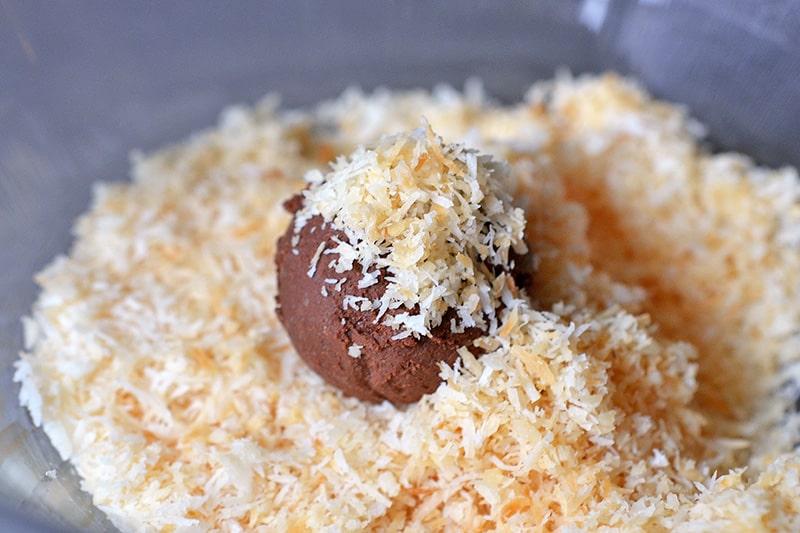 A Nom Nom Paleo Chocolate Truffle is in a bowl of toasted shredded coconut.