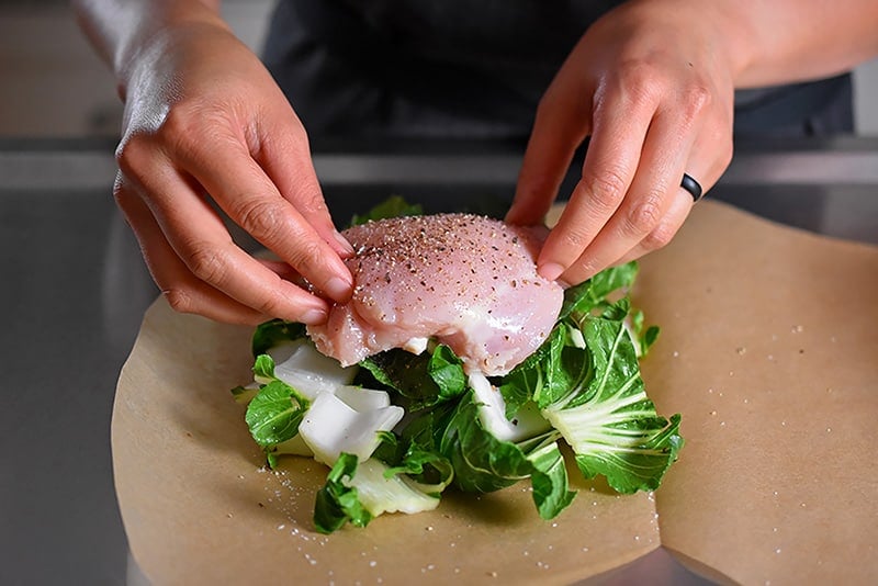 Someone placing the raw and seasoned chicken on top of the bok choy for the paper-wrapped chicken recipe.