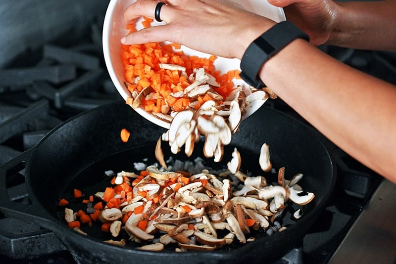 Adding carrots and mushrooms into a large cast iron skillet.