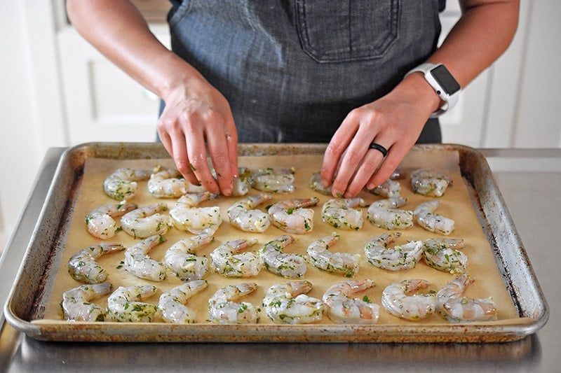 Someone placing the marinated shrimp in rows on a parchment lined baking sheet.