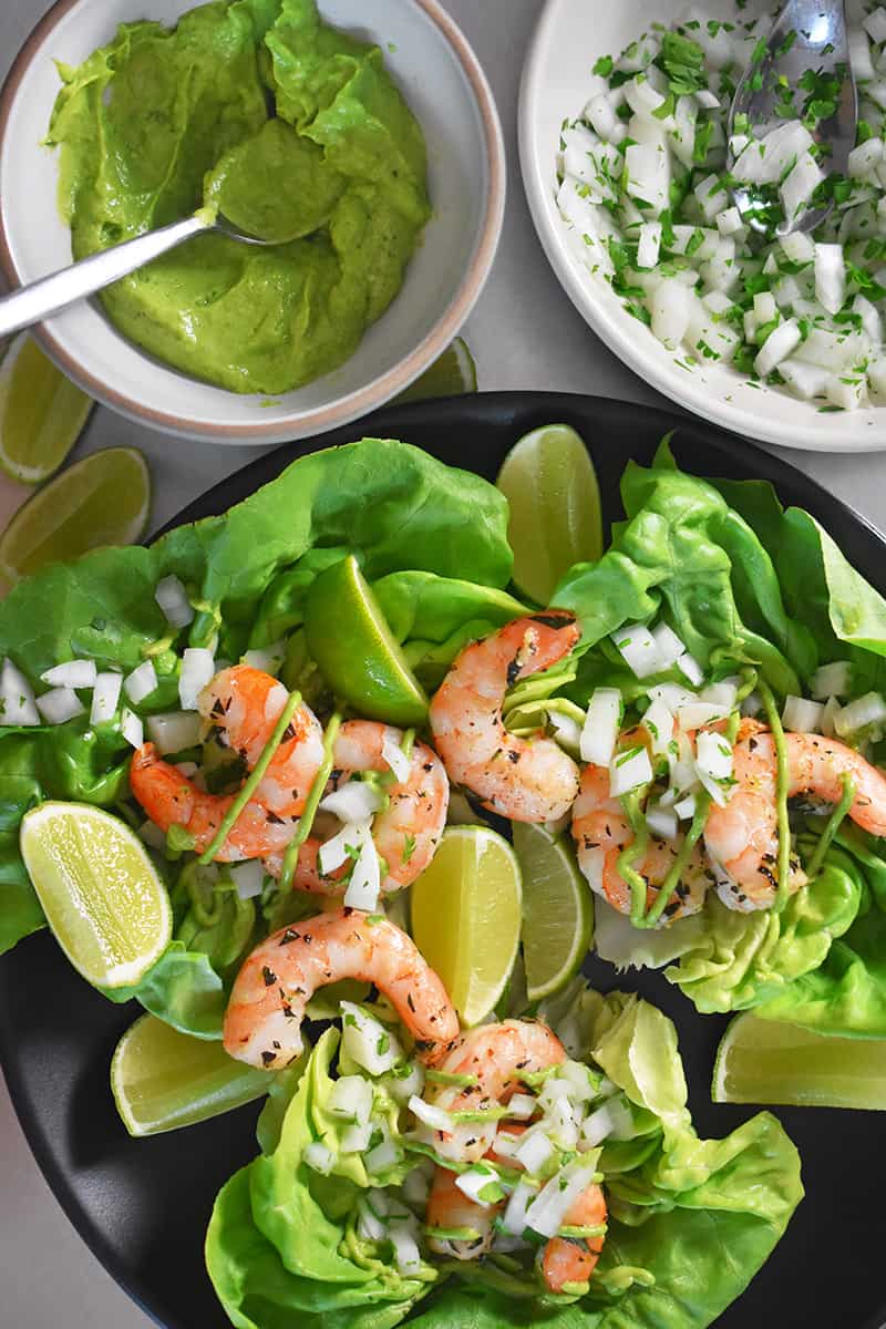 Paleo shrimp tacos in a bed of lettuce sitting on a plate with slices of lime. Surrounding the plate is a bowl of guacamole and a bowl of chopped onions.