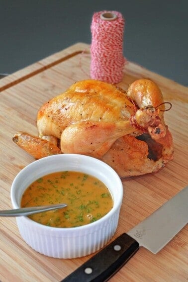 A closeup shot of Weeknight Roast Chicken on a wooden cutting board with gravy in a ramekin.